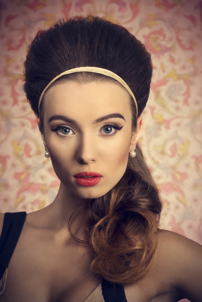 Short portrait of young, beautiful woman in fitted cream and black dress is looking down. She has got big blue eyes, brown big hairbun and she is wearing hairband and pearls on her ears.