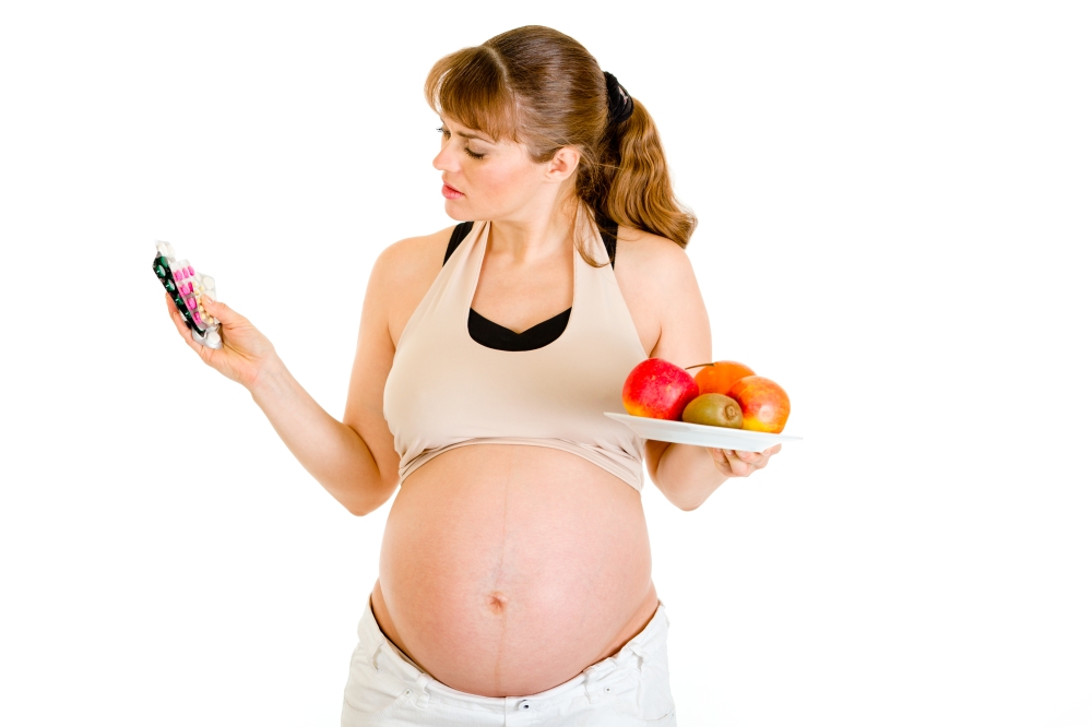 Pensive pregnant woman making choice between drugs and fruits isolated on white&#xA;