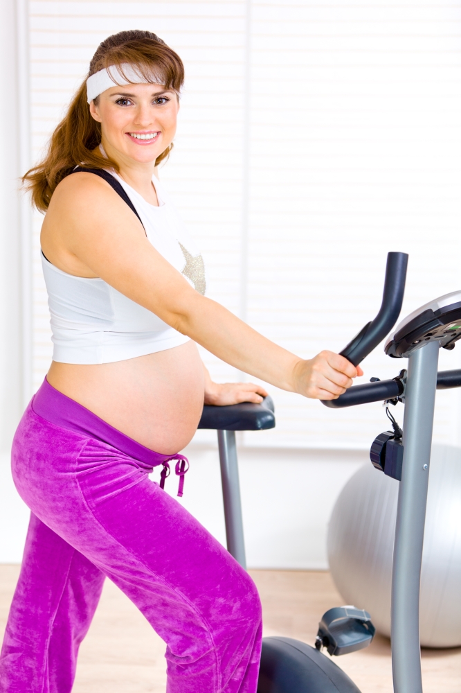 Smiling beautiful pregnant woman preparing for workout on static bicycle&#xA;