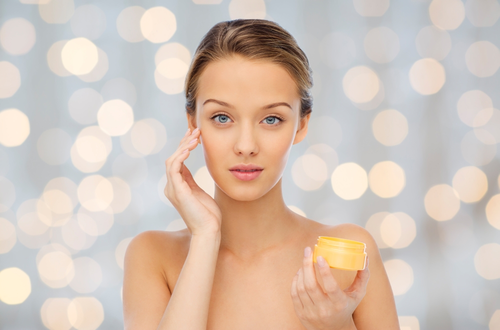 beauty, people, cosmetics, skincare and cosmetics concept - young woman applying cream to her face over holidays lights background
