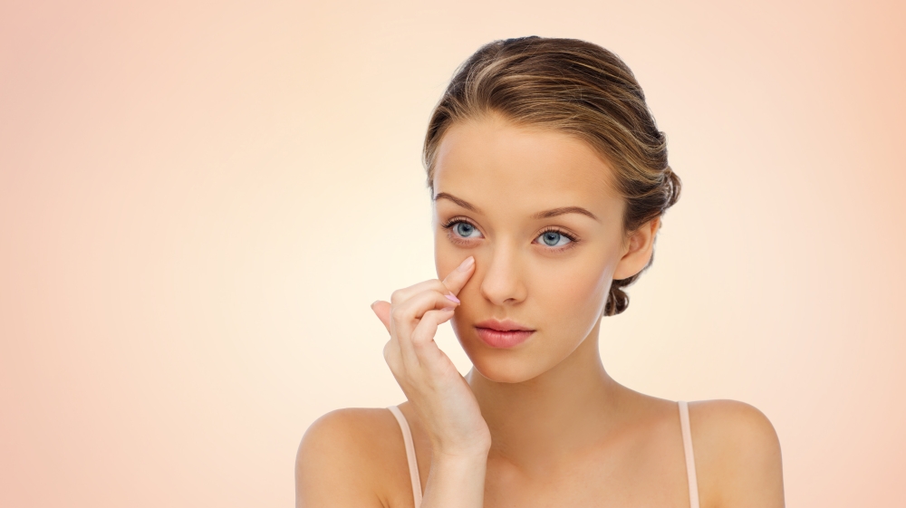 beauty, people, cosmetics, skincare and health concept - young woman applying cream to her face over beige background