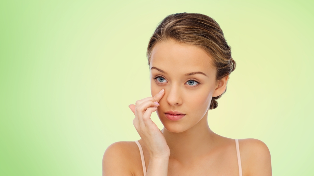 beauty, people, cosmetics, skincare and health concept - young woman applying cream to her face over green background