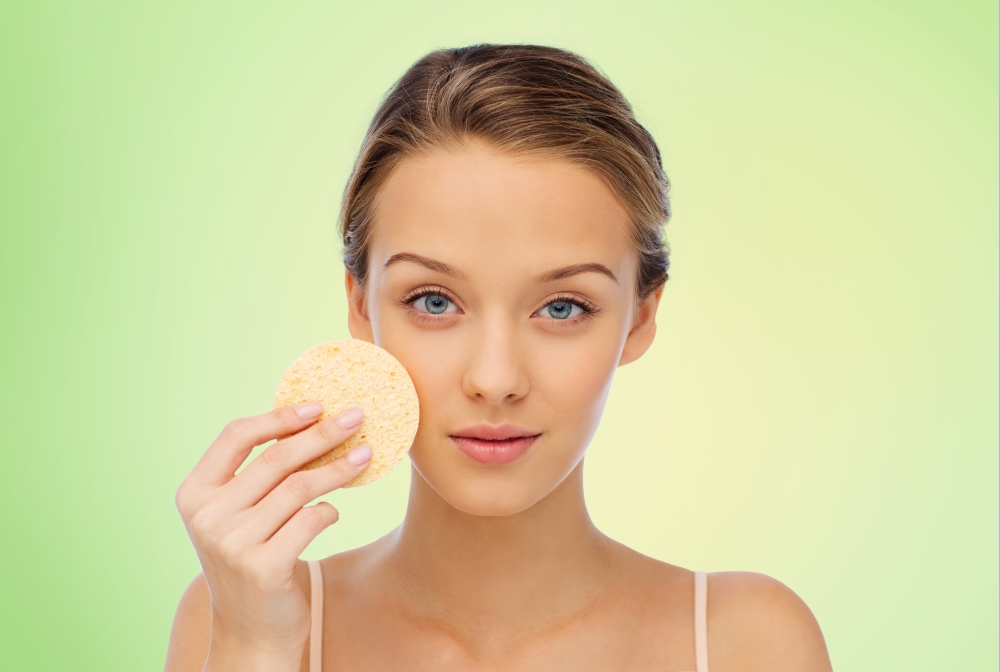 beauty, people and skincare concept - young woman cleaning face with exfoliating sponge over green background