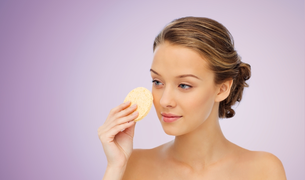 beauty, people and skincare concept - young woman cleaning face with exfoliating sponge over violet background