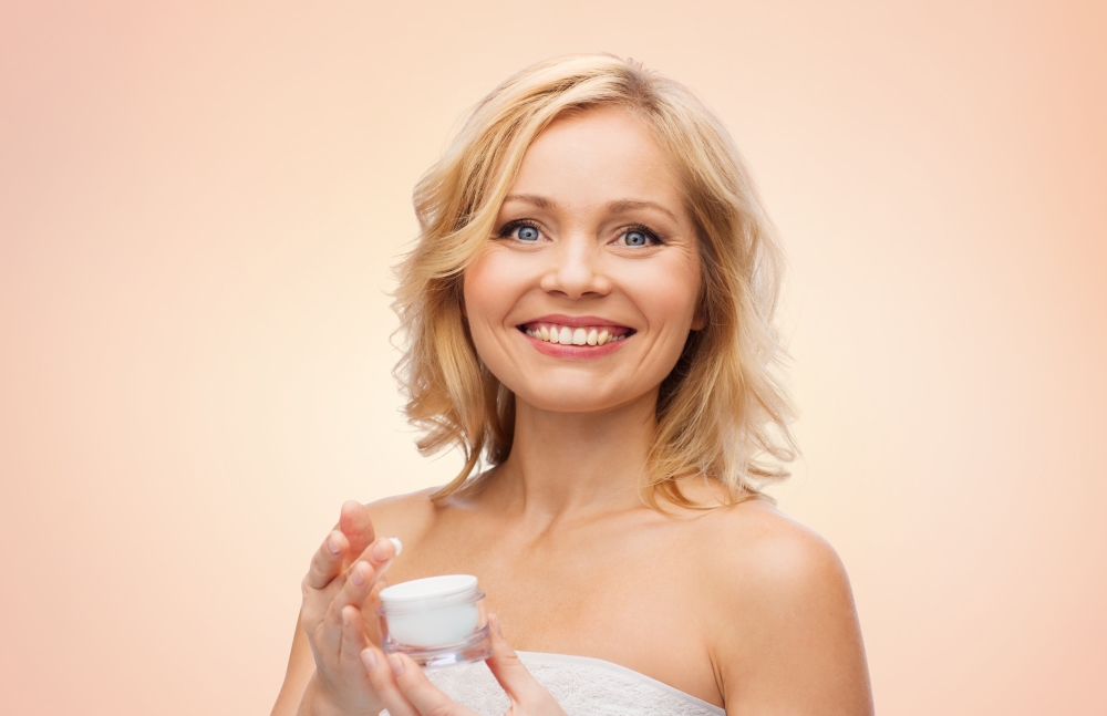 beauty, people, skincare and cosmetics concept - happy middle aged woman with cream jar over beige background