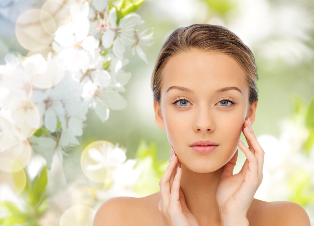 beauty, people, summer, organic and health concept - young woman with bare shoulders touching her face over green natural background with cherry blossom