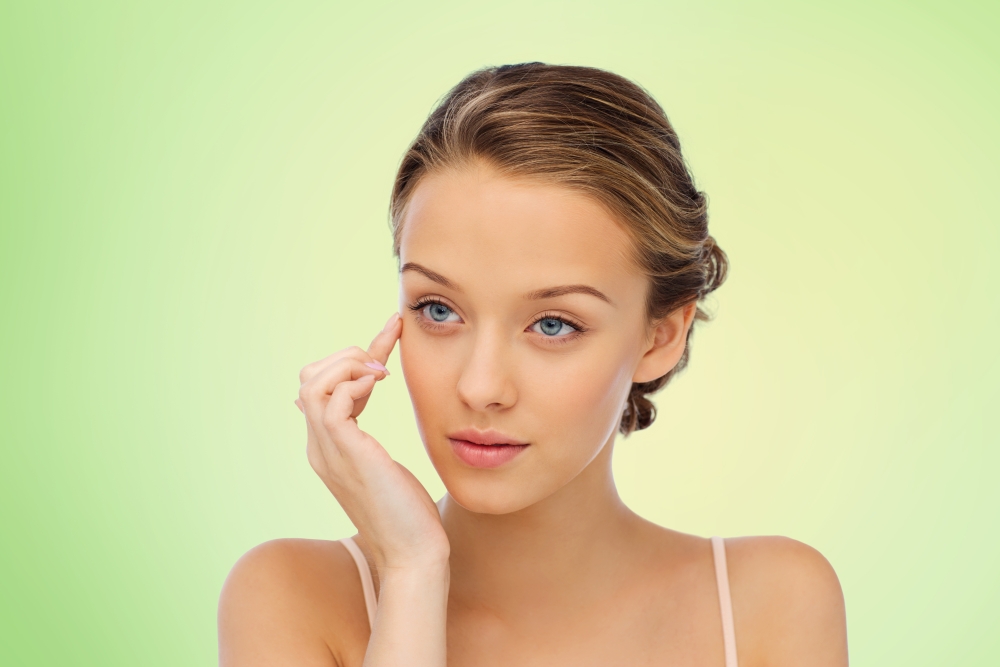 beauty, people, cosmetics, skincare and health concept - young woman applying cream to her face over green natural background