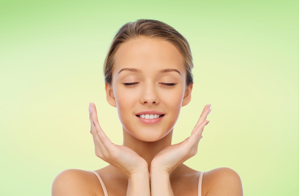 beauty, people, skincare and health concept - smiling young woman face and hands over green natural background