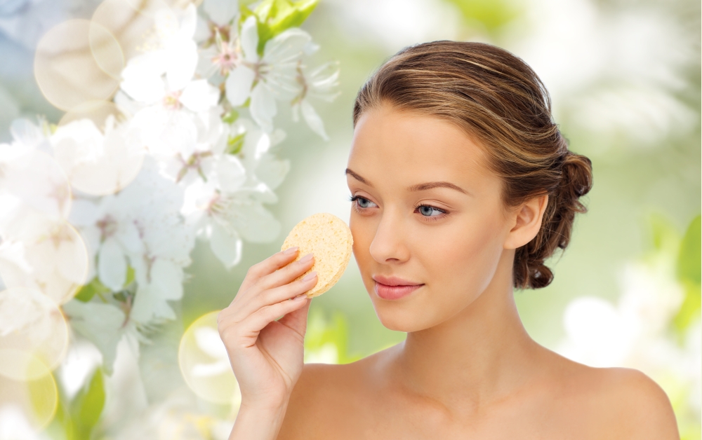 beauty, people and skincare concept - young woman cleaning face with exfoliating sponge over green natural cherry blossom background