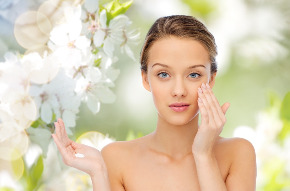 beauty, people, cosmetics, skincare and health concept - happy smiling young woman applying cream to her face over green natural cherry blossom background