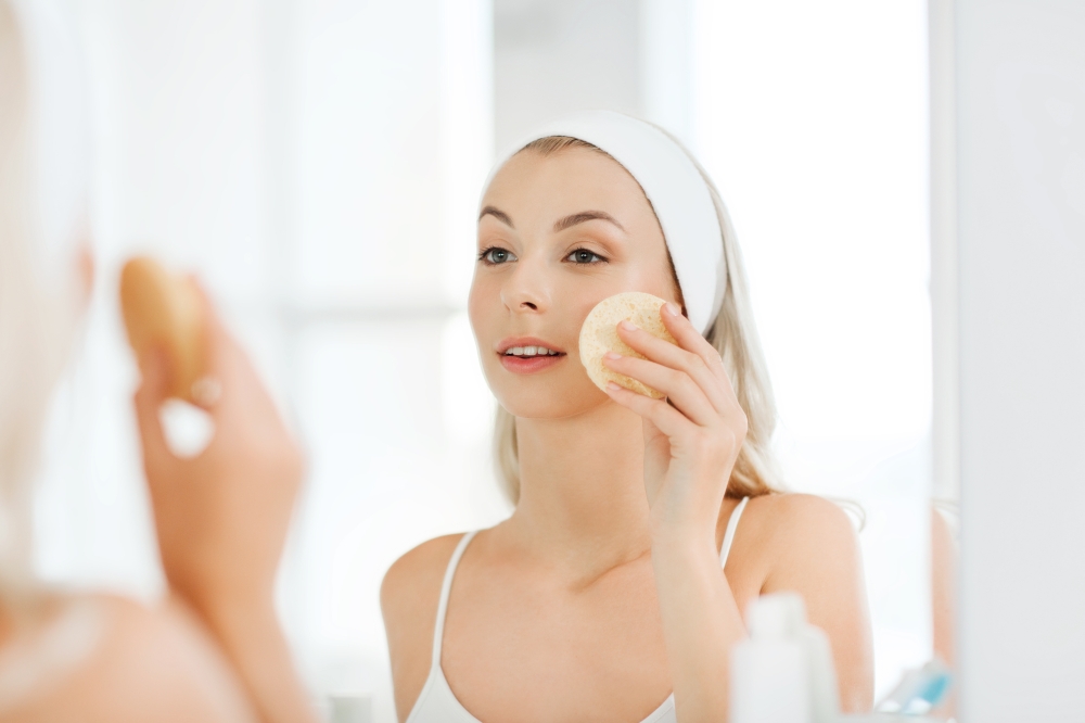 beauty, skin care and people concept - smiling young woman washing her face with facial cleansing sponge at bathroom