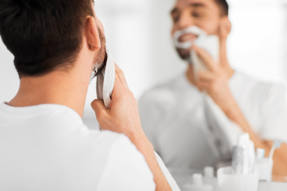 beauty, shaving, grooming and people concept - close up of man removing shaving foam from face with towel and looking to mirror at home bathroom