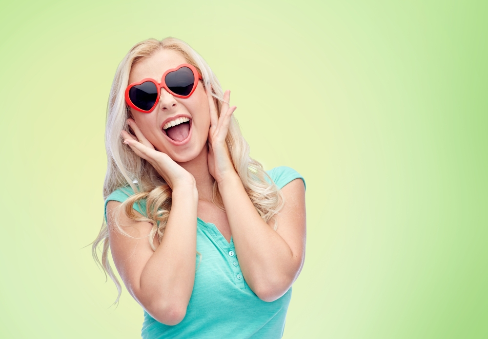 emotions, expressions, summer and people concept - smiling young woman or teenage girl in heart shape sunglasses over green natural background