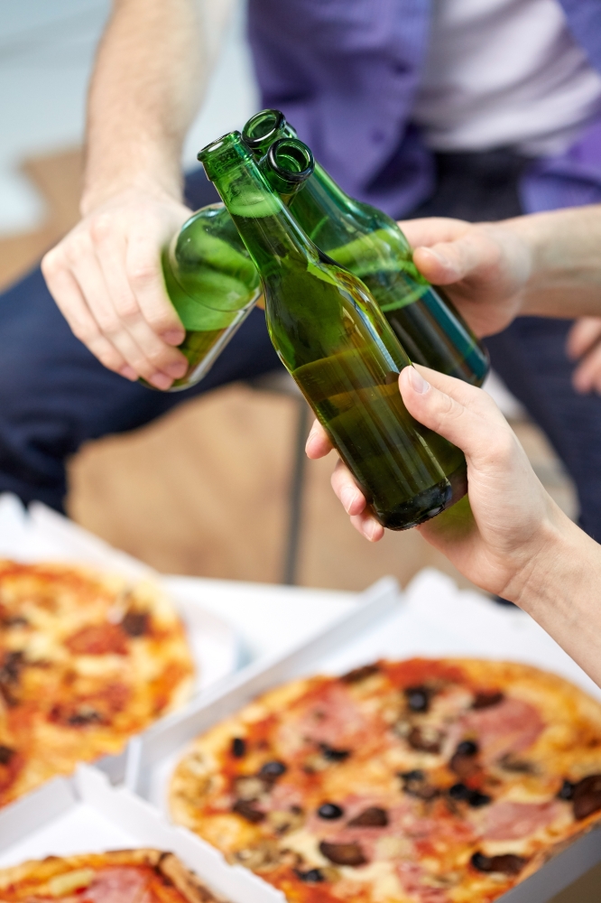 friendship, alcohol, people, celebration and holidays concept - close up of male hands clinking beer bottles and eating pizza at home