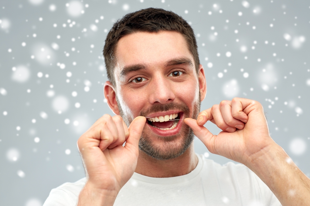 dental hygiene, care, people and winter concept - smiling young man with floss cleaning teeth over snow on gray background