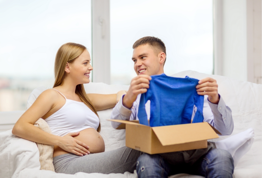 pregnancy, post, delivery and parenthood concept - happy family expecting child sitting on sofa and opening parcel box with blue cardigan