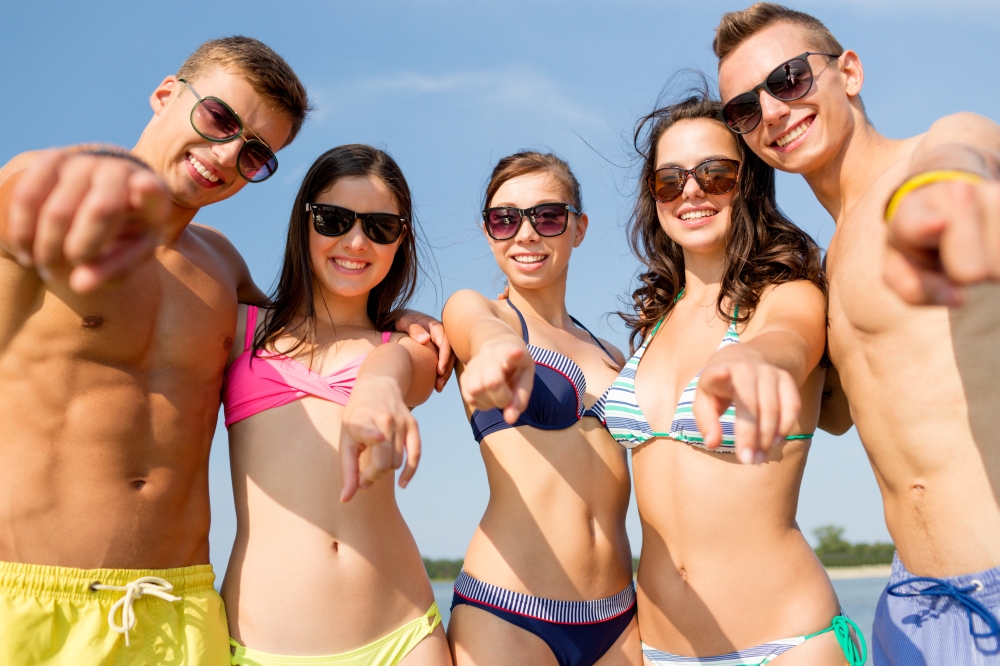 friendship, sea, holidays, gesture and people concept - group of smiling friends wearing swimwear and sunglasses pointing on you on beach