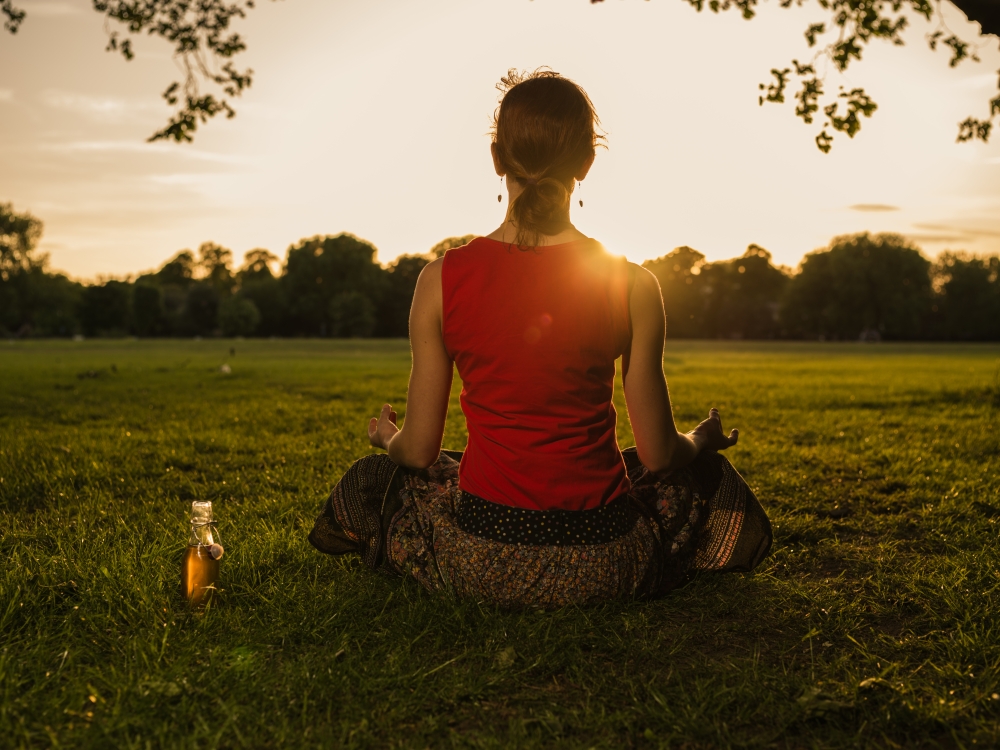 A young woman is meditating in a park at sunset, there is a bottle of health drink next to her