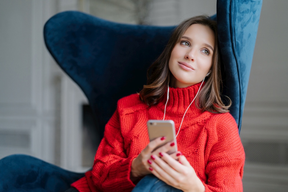Thoughtful woman in a red turtleneck listening to music on her phone, relaxed in a high-back blue chair, daydreaming