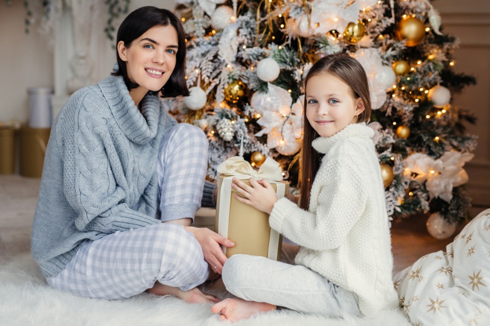 Mother and daughter with Christmas gift by a festive tree in cozy attire