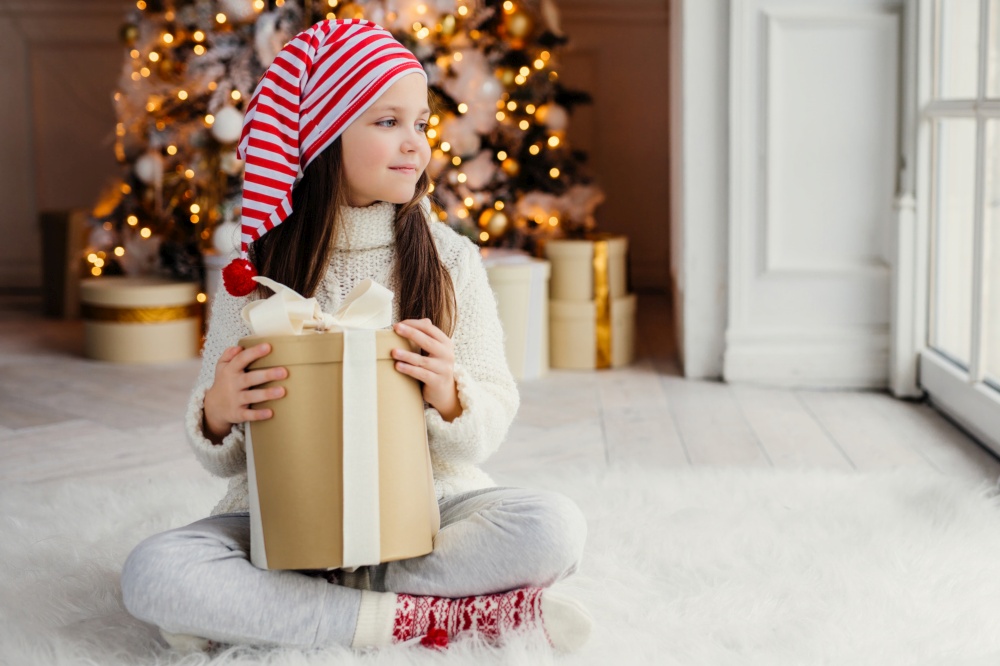 Girl in festive hat holding a Christmas gift, tree with golden lights behind