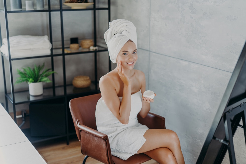 Joyful woman with a towel turban applying face cream, in a chic interior