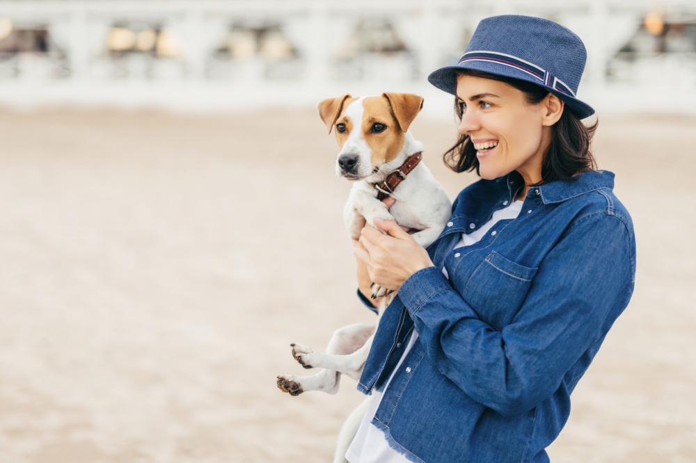 Joyful woman in a blue denim shirt and fedora holding her beloved Jack Russell Terrier on the beach, both looking playfully towards the sea
