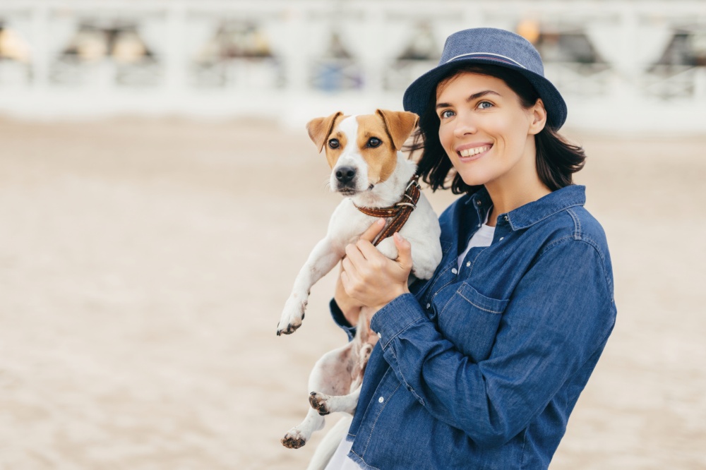 Cheerful woman in blue attire holding a playful Jack Russell Terrier on a sandy beach, both looking ahead with bright, happy eyes