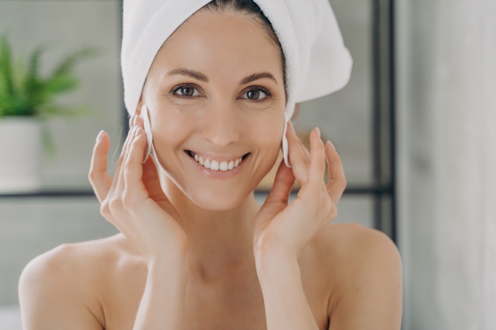 Radiant woman with a towel on her head gently touching her face with a cotton pad, showcasing a daily skincare routine