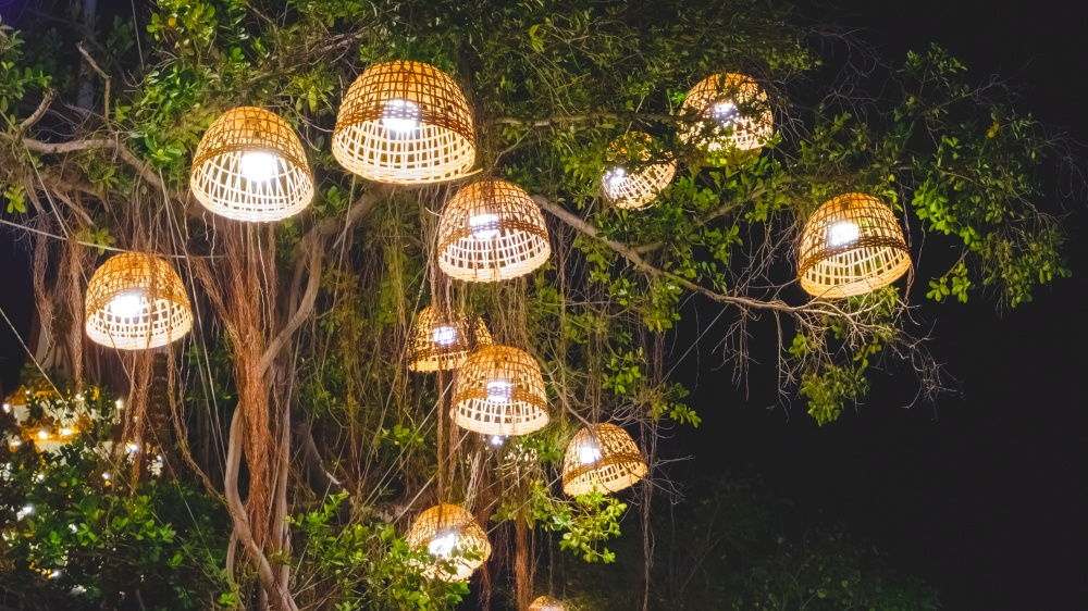 Decorative illuminated lighting of many vintage bamboo lamps with chicken coops hanging on trees in outdoors event area at night garden