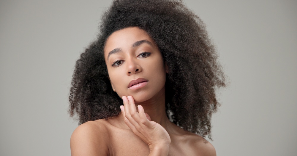 Beauty and healthcare concept - beautiful African American woman with curly afro hairstyle and clean, healthy skin touches her cheek and face with her hand, posing and looking at the camera. High quality photo. Beauty and healthcare concept - beautiful African American woman with curly afro hairstyle and clean, healthy skin touches her cheek and face with her hand, posing and looking at the camera