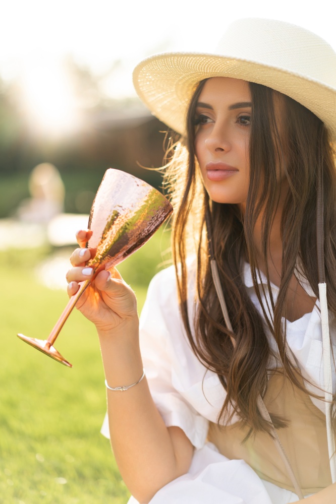 beautiful glamorous girl in a white dress and a straw hat on the shore of the lake with a goblet in her hands