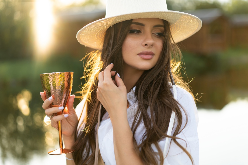 beautiful glamorous girl in a white dress and a straw hat on the shore of the lake with a goblet in her hands