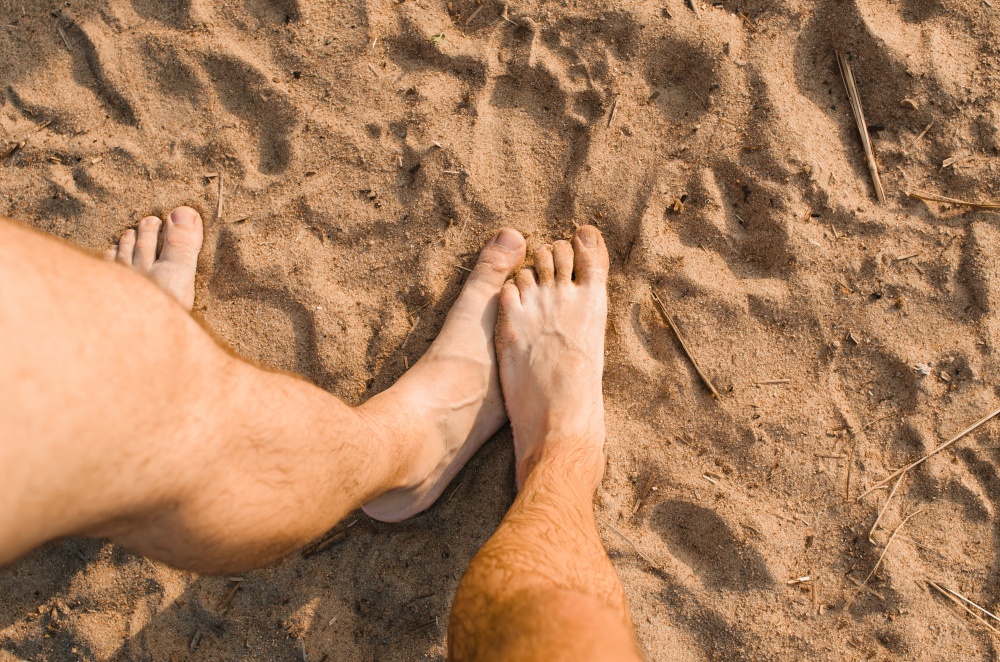 Homosexual relationship concept. Male hairy foot touching other male foot on the beach, top view. Hidden touching each other while relaxing outside.