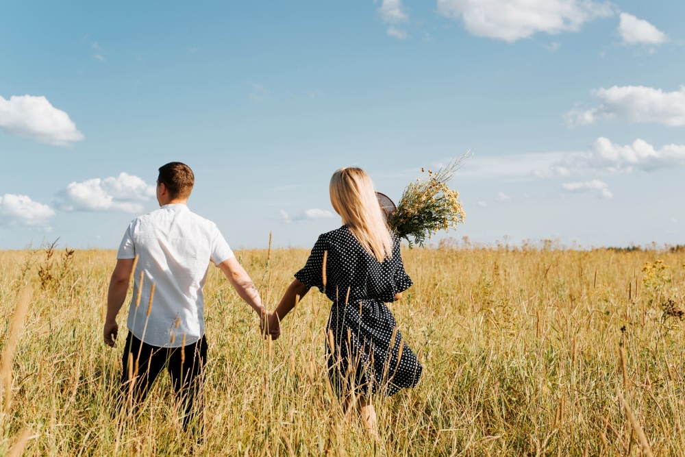 Happy young couple holding hands and walking in field, boyfriend and girlfriend in summer dress with bouquet of wild flowers, back view. Beautiful people in love enjoying time together. Love concept.. Happy young couple holding hands and walking in field, boyfriend and girlfriend in summer dress with bouquet of wild flowers, back view. Beautiful people in love enjoying time together. Love concept