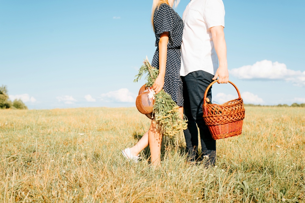 Romantic picnic in nature. Cropped image of young couple hugging in field on meadow, man holding picnic basket, young woman in summer dress holding bouquet of wildflowers and straw hat. Love story.. Romantic picnic in nature. Cropped image of young couple hugging in field on meadow, man holding picnic basket, young woman in summer dress holding bouquet of wildflowers and straw hat. Love story