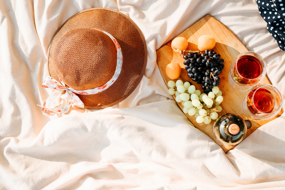 Picnic in nature. Top view of bottle of red wine, two glasses of alcohol, grapes, peaches on wooden board, straw hat on picnic blanket. Recreation, dating, outdoor leisure concept