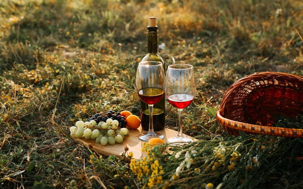 Romantic date picnic outdoors, still life. Bottle of red wine, two glasses, fruits, grapes, peaches, wicker basket in field at sunset outdoors.. Romantic date picnic outdoors, still life. Bottle of red wine, two glasses, fruits, grapes, peaches, wicker basket in field at sunset outdoors