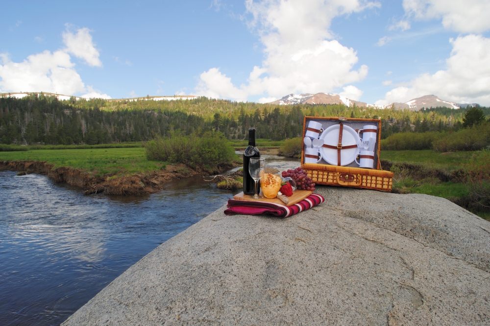 Fruit and Wine picnic in a scenic mountain setting with a meadow and river in the background. Picnic On The River