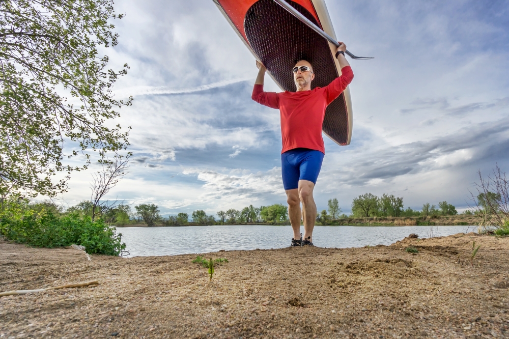 senior male paddler carrying his SUP paddleboard on a lake shore in Colorado, early spring