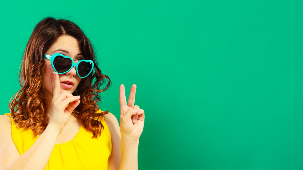 Summer fashion eyes protection concept. Closeup girl long curly hair in heart shaped sunglasses making thumb up hand sign on green vivid color background